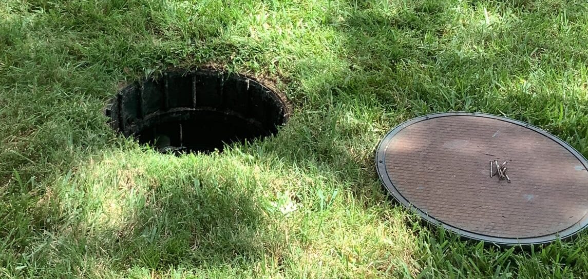 A septic lid lying on the ground next to a hole leading into a septic tank.
