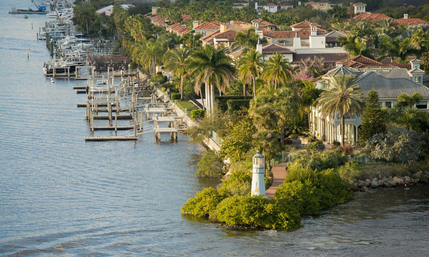 Waterfront luxury homes in Florida with palm trees, private docks, and a small lighthouse on the shoreline, overlooking calm waters.